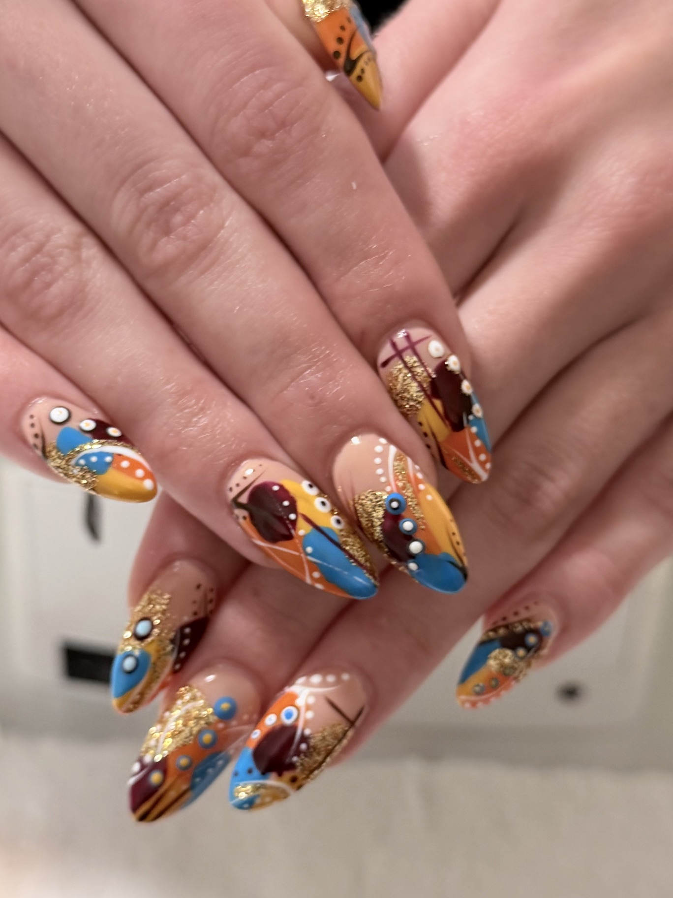 Nude almond nails with abstract orange, blue and burgundy shapes, white dots and gold glitter